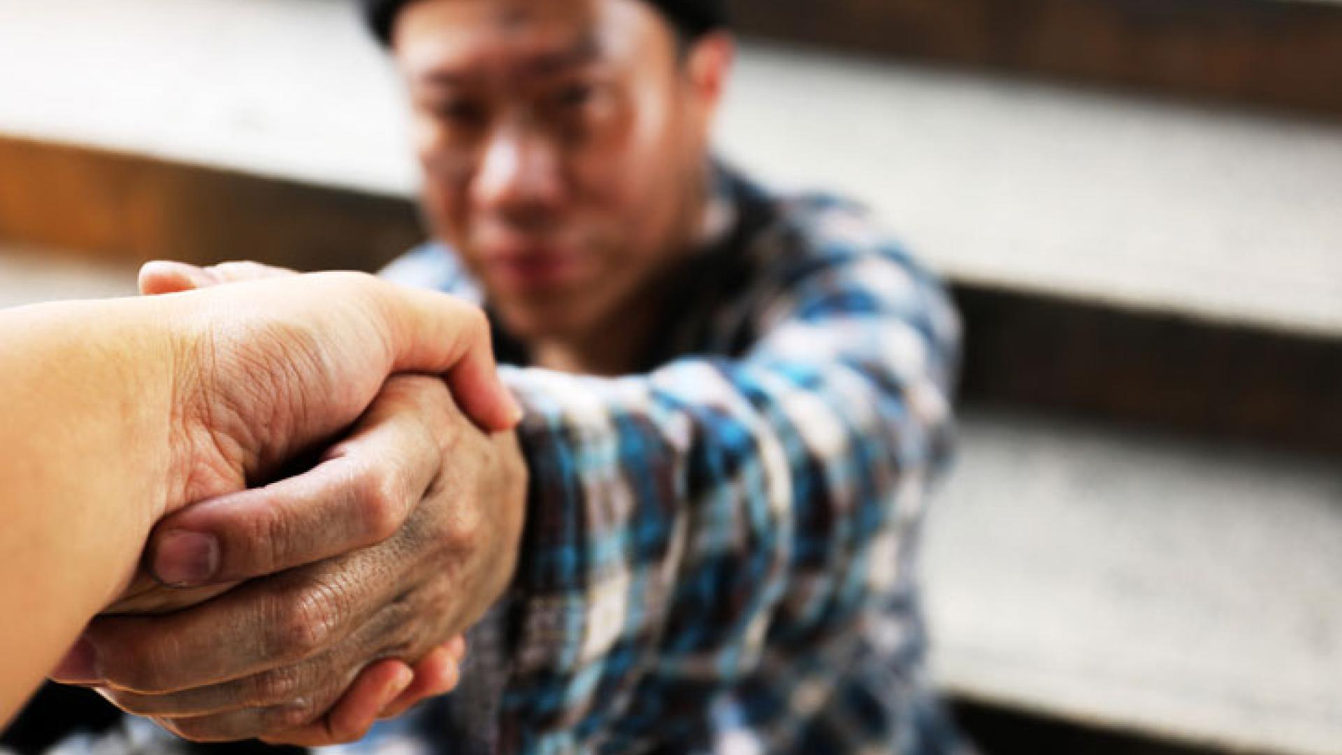An outstretched hand in the left foreground holds the hand of a slightly-blurry asian man, who is sitting down and wearing a blue plaid shirt and black tuque.