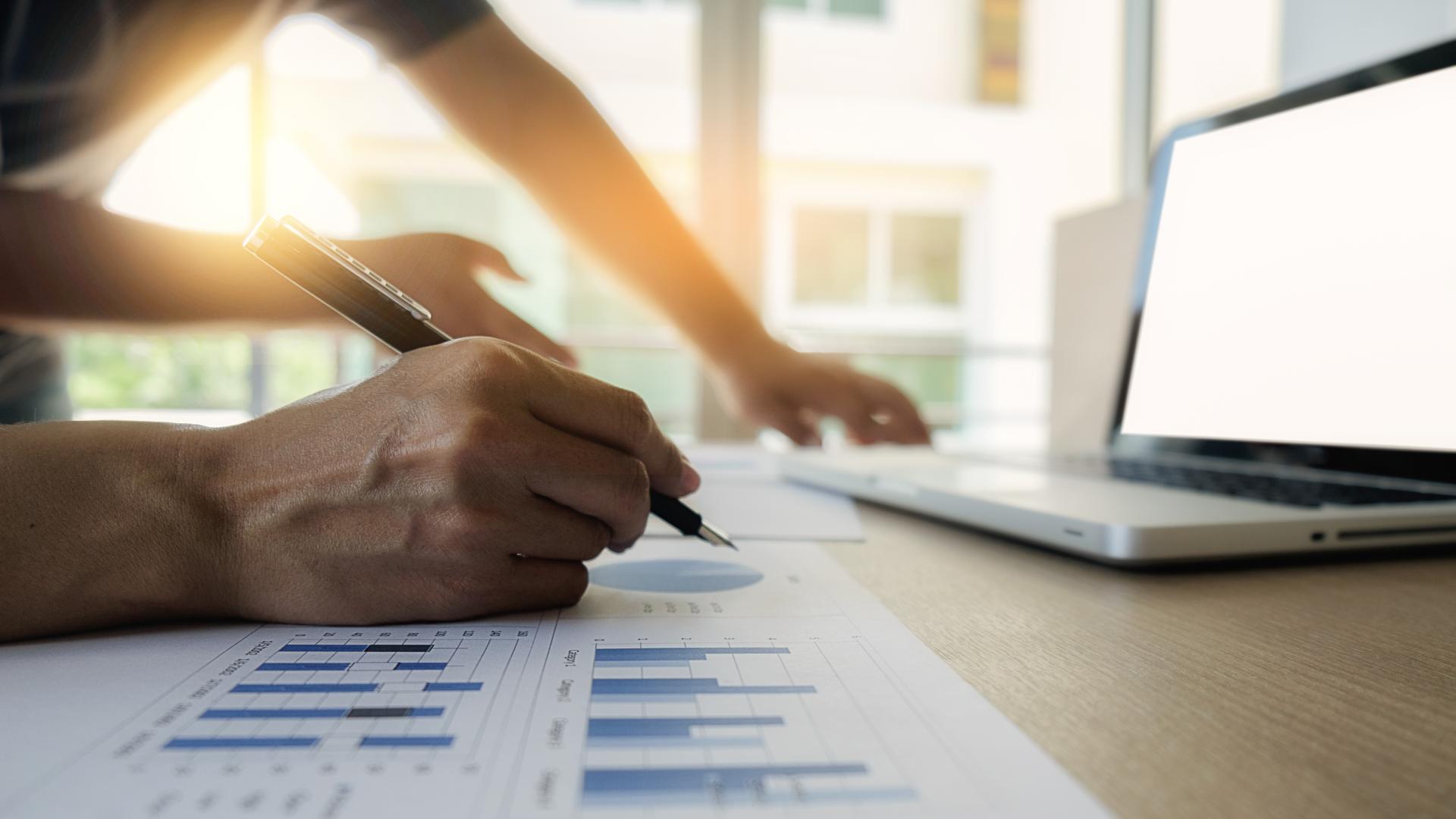 Close-up of hands working on graphs with a pen and laptop, indicating analysis and teamwork in a professional setting